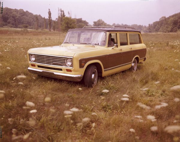 Three-quarter view from front left of a man sitting in the driver's seat of a 100 Travelall parked in a field. The Travelall exterior is yellow with woodgrain panels.