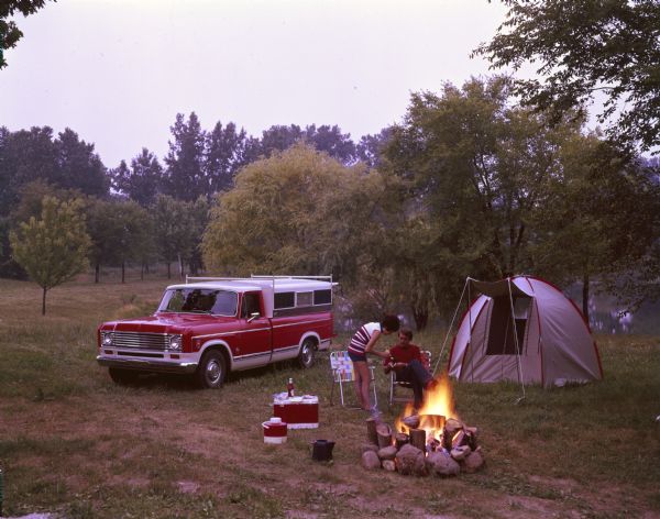 A man and woman are eating in front of a campfire. A red 100 pickup is parked on the left. In the background is a tent. The campsite is set up on the shoreline of a river or lake.