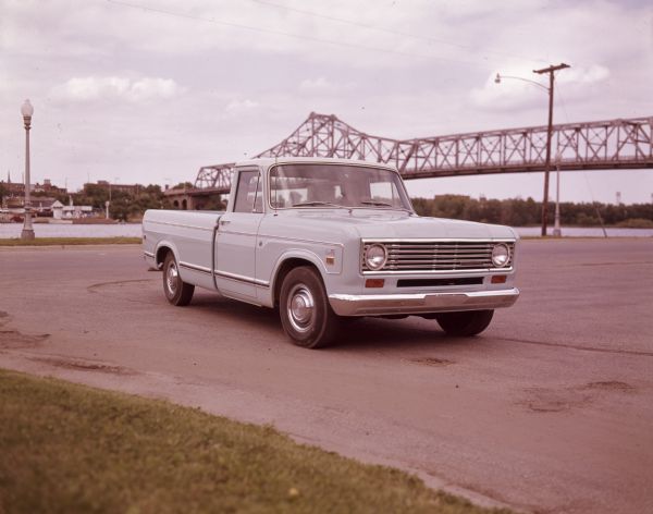 Three-quarter view from front right of a man sitting in the driver's seat of a 100 Special Pickup. The truck is parked outdoors in a parking area with a bridge over water in the background.