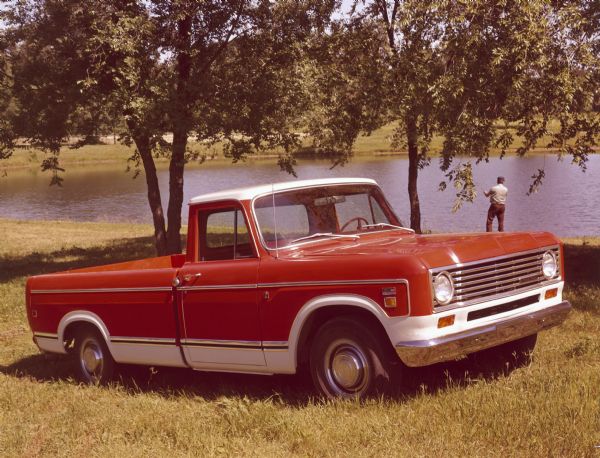 Three-quarter view from front right of two tone, (flame red with white) pickup with open truck bed parked in the grass. In the background a man is fishing along the tree-lined bank of a body of water.