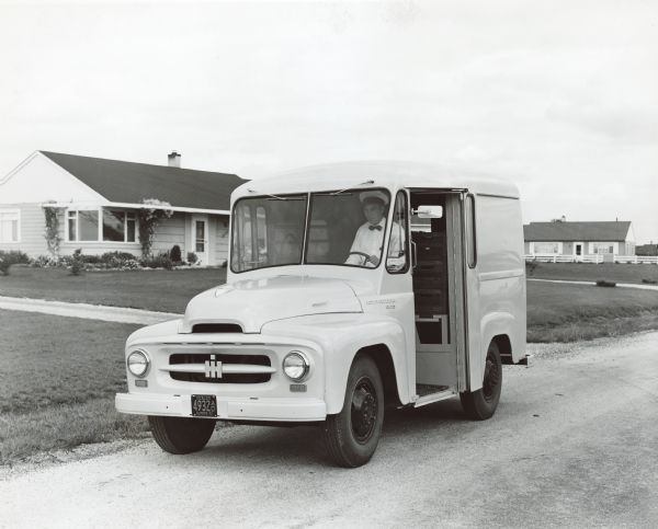 Three-quarter view from front of driver's side of a RBA-140 Milk Delivery Truck. A man, wearing a white uniform with a hat and a bow tie, is sitting in the driver's seat with the door open. The truck is on parked on a road, and in the background are houses.