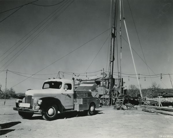 Three-quarter view from front left of a KB-3 truck with specially built body to carry 300 quarts of nitroglycerine. The truck is being loaded at the dump, which is located a safe distance from any buildings. A sign on the side of the truck reads: "Nitro." On the front is a sign reading: "Explosives." Two men are standing on the platform of an oil derrick behind the truck.