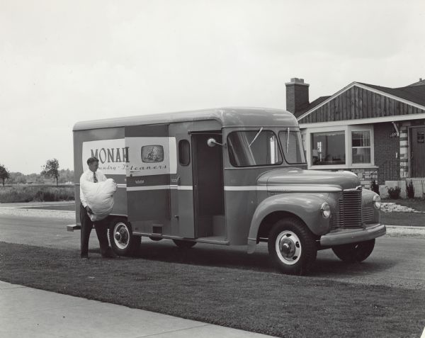 View from sidewalk of a man holding a bag of laundry standing near the partially open side door of an International KB-5 truck parked at a curb. The truck has a special body designed by Monarch Laundry. On the other side of the street is a house.