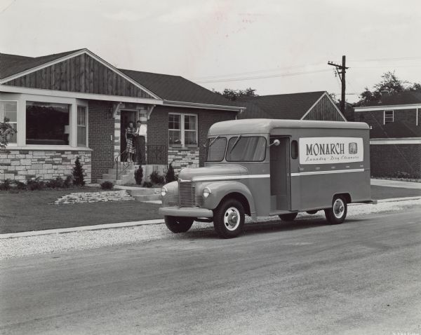 View across street towards a an International KB-5 truck parked along a curb. Behind the truck is man talking to a woman at the open front door of a house. The truck has a special body designed by Monarch Laundry.