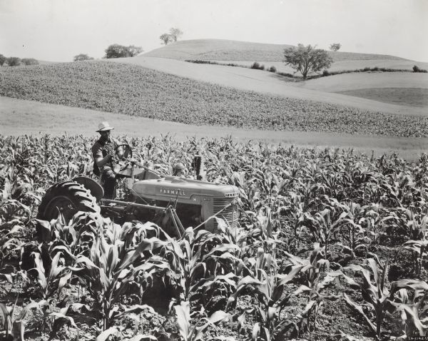 Three-quarter view from front right of a man cultivating corn in stripped-cropped field using a Farmall H tractor and HM-238 Farmall cultivator. In the background are hills and trees. F.F. Miller farm five miles west of West Salem, Wisconsin.