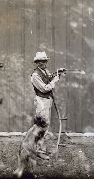 A man is standing in front of a barn holding a scythe while sharpening it. A dog is in the foreground.
