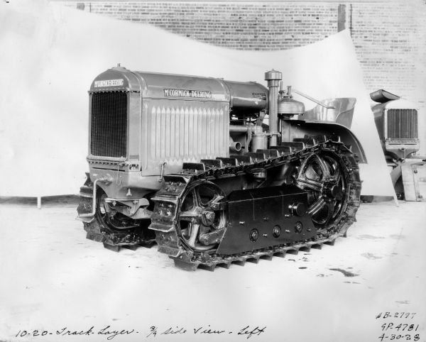 Three-quarter view from front left of a McCormick-Deering 10-20 Tractractor. In the background a white sheet has been hung as a backdrop, and part of a brick wall is above the sheet. Part of another tractor is in the background on the right.