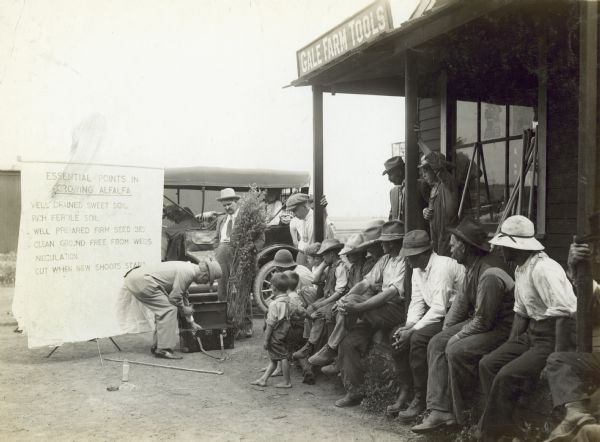 Men seated near public speaker demonstrating proper cultivation of alfalfa. Original caption reads: "This scene was taken during the Alfalfa Campaign in Dupage County. Showing the speaker picking out the samples of sweet and sour soil, of limestone, rock phosphate, litmus paper, and other things. This is one of the cases carried with the speaker for the purpose of further demonstrating to the crowd the methods of feeding alfalfa."