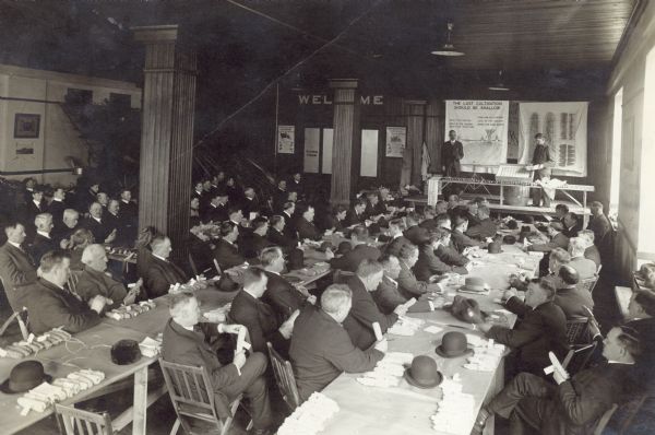 Elevated view of a group of men attending an Agricultural short course in Madison, Wisconsin. There are samples of ears of corn on the tables. Two men stand at the end of the room on a platform. Behind them one of the large posters reads: "The Last Cultivation Should Be Shallow." 87 IHC employees, consisting of General Agents, Asst. Gen. Agents, Blockmen, and Travelers were present.
