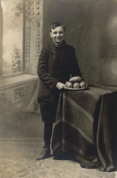 Full-length studio portrait of a boy, 14-years-old, posing in front of a painted backdrop. He is standing next to a cloth covered table holding a plate of potatoes.