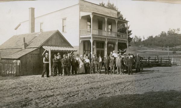 Group portrait of the Bliss Alfafa Club of Emmet County, showing the President Mr. Grant. The group of men, women and children is standing on an unpaved road with in front of two buildings. Children are standing in the back of an automobile parked behind the group.