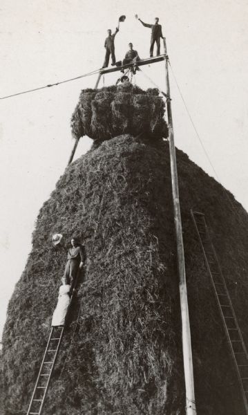 Men posing on top of a tall stack of alfalfa. The derrick is homemade. A man and a woman are posing on a ladder against the stack of alfalfa.