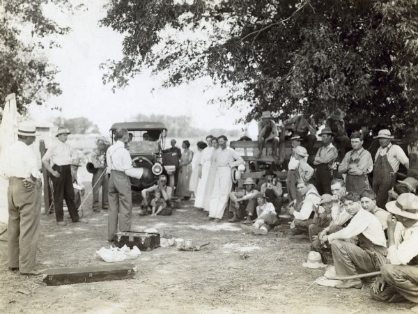 Group of men, women and children gather outside around a man lecturing about growing alfalfa.