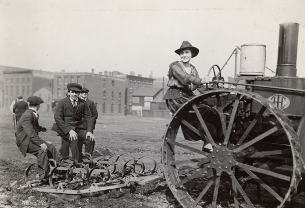 Miss Josephine Huddleston seated on the big IHC tractor at the plowing of the first tract of land at Harrison and Jefferson Streets for Chicago's City Garden Campaign. A group of young boys are sitting on the plow. Brick buildings are in the background.