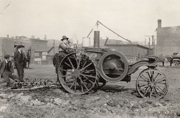 Miss Josephine Huddleston seated on the big IHC tractor at the plowing of the first tract of land at Harrison and Jefferson Streets for Chicago's City Garden Campaign. A man is standing behind Miss Huddleston. A group of young boys are sitting and standing on the plow. Brick buildings are in the background.