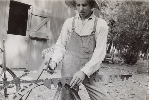 Close-up of a young man removing sections from a sickle with a hammer. There is a barn in the background.