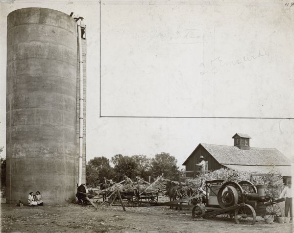 View across barnyard towards men loading corn into a silo on a farm. The men are working on the right with horse-drawn wagons loaded with cornstalks. A tractor is belt-driving machinery that is blowing the silage up and into the open top of the silo. A man is standing at the top of the silo. Three young girls and a dog are sitting at the base of the silo on the left.