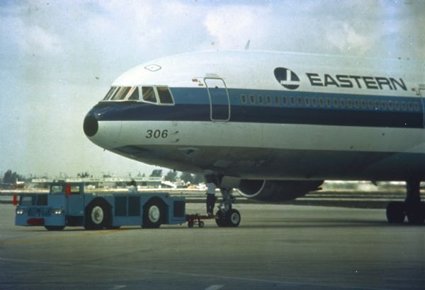 View across tarmac towards a man using a Hough T-500 Paymover  an airport pulling a jet airliner operated by Eastern Airlines.
