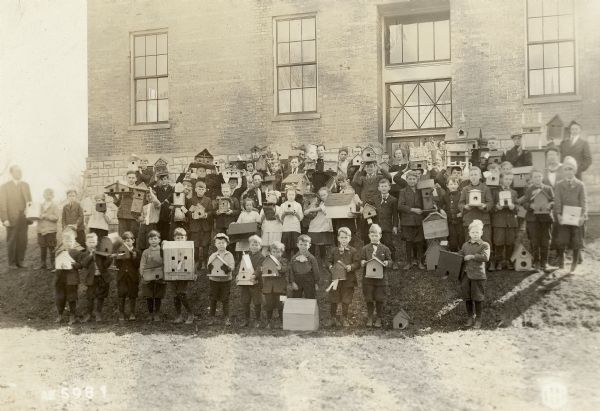 Group portrait of school children posing outdoors on a slope in front of a school building. The children are all holding birdhouses, probably handmade. Location unknown.
