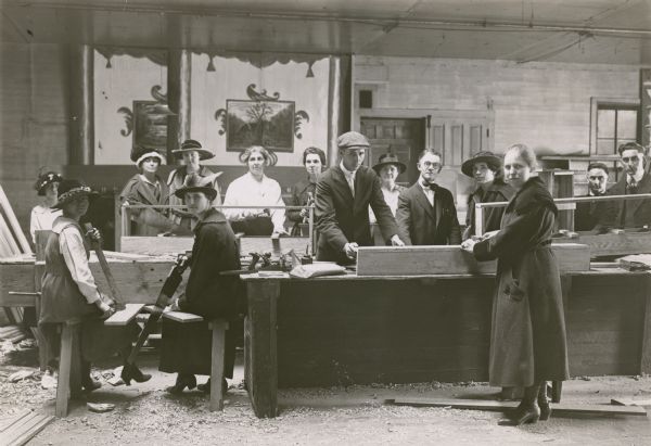 Group portrait of Mercer County teachers, men and women, making a project in vitalized agriculture. They are in a wood workshop, and some of the women are holding saws, hammers and wood boards. Other men and women are posing along the workbenches.