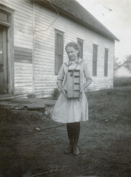 Outdoor portrait of Edna Baker, first prize girl, standing and holding a nail box. Behind her is probably the Pleasant Valley School house in Logan County.