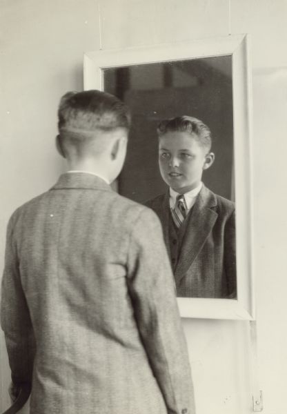 Portrait of young man, wearing a suit and tie, standing in front of a mirror on the wall looking at his reflection.