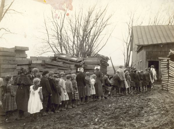 Group of children walking on wooden boards to get to an onion shed. There is mud in the driveway. Pallets are stacked on the left and right.