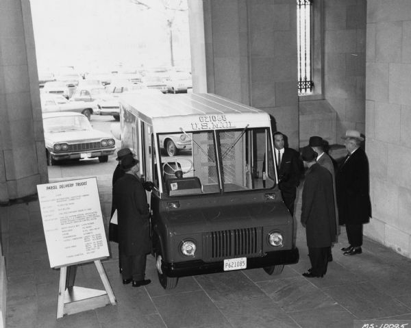 Elevated view of an International M-800 Metro. Six men surround the vehicle that is parked inside a covered alcove. Cars are seen parked in the background. Original caption reads, "Post office department officials in Washington D.C., look over the display of a new International M-800 Metro. A total of 2,806 of these forward-control multi-stop compacts currently are being delivered to post office facilities throughout the United States. Cost of the units, which will be used for regular mail and parcel post delivery, was $6.5 million. This was the largest order for multi-stops International has ever received. The trucks are equipped with right-hand drive and automatic transmissions."