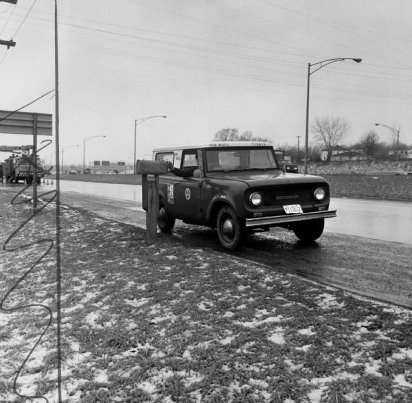 A three-quarter view of a International scout postal truck. A mail carrier is reaching out the left-hand window and placing mail in a mail box.  
