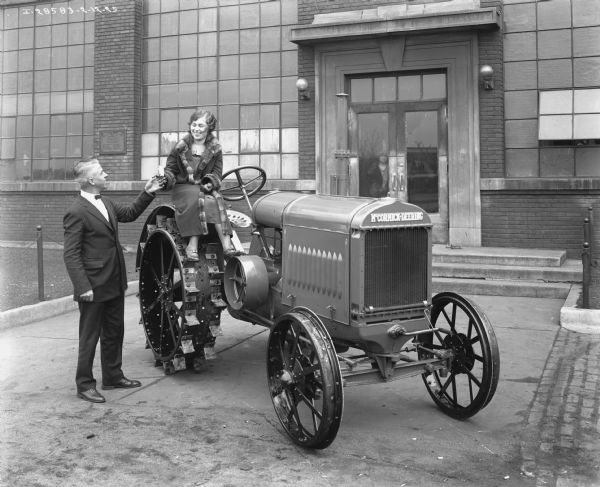Outdoor portrait in front of an International Harvester building of a man wearing a suit and bow tie standing next to a woman wearing a fur-trimmed coat. The woman is sitting on the fender over the right wheel of a McCormick-Deering tractor. The woman is holding a small bag in her hand, and the man is reaching up his hand to hers. The tractor is parked in front of the entrance to the building, and a man and woman are standing inside the glass doors looking out.