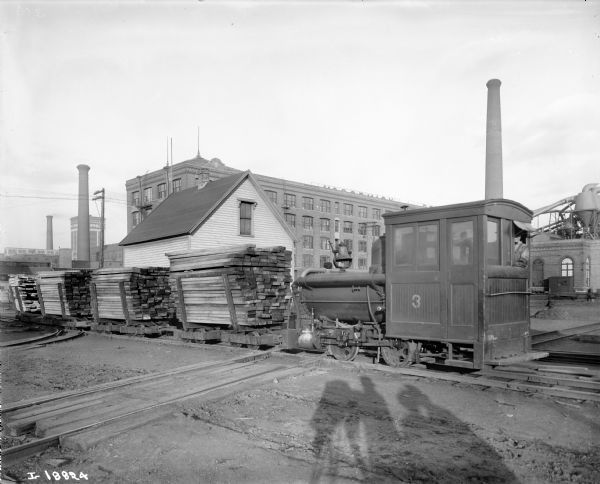 Railroad yard at McCormick Works. A man is looking out the window of the locomotive on the right, and there are stacks of lumber on the small, open railroad cars. The shadow of the photographer, his camera and two other people can be seen in the foreground. Factory buildings and smokestacks are in the background.