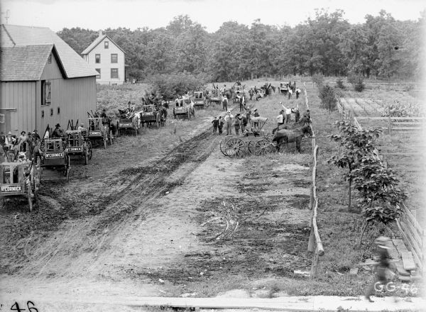 Elevated view of a parade of farmers in horse-drawn wagons picking up their new McCormick machinery packed in crates. They are parked along a rural dirt road on the outskirts of town. Known as "McCormick Days," such events were organized by local dealers to advertise McCormick machinery and implements after they arrived from shipment. A number of the men are raising their hats for the group portrait.