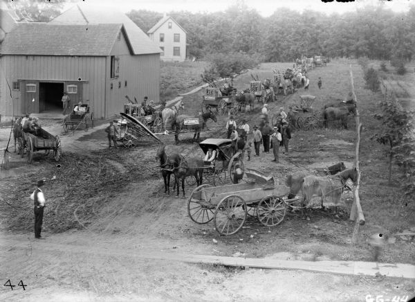 Elevated view of a parade of farmers and young children with horse-drawn wagons picking up their new McCormick machinery. They are parked along a rural dirt road on the outskirts of town. Known as "McCormick Days," such events were organized by local dealers to advertise McCormick machinery and implements after they arrived from shipment. A corn binder is near the barn on the left.
