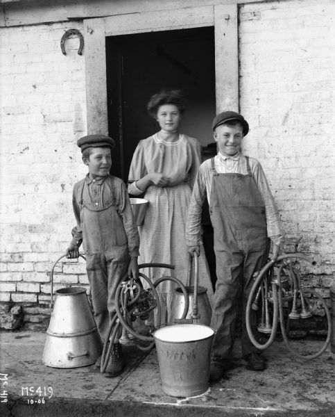 Two boys dressed in dark work shoes, overalls, and button-up shirts are standing on either side of a woman in front of a doorway. The woman is dressed in a long skirt and blouse, and has her hair pulled into a bun on top of her head. All three of them are smiling, and holding buckets or milk cans. The boy on the right is holding milking machinery. Behind them is a whitewashed brick building, and a horseshoe hangs at the top left of the doorway.