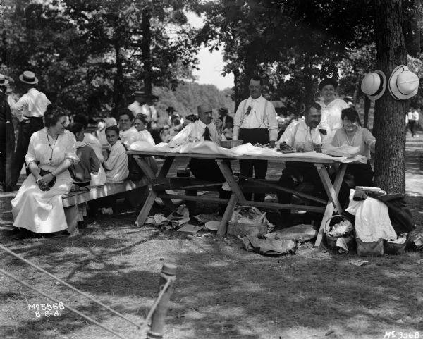 View of men, women and children sitting on benches near picnic tables set out under trees.