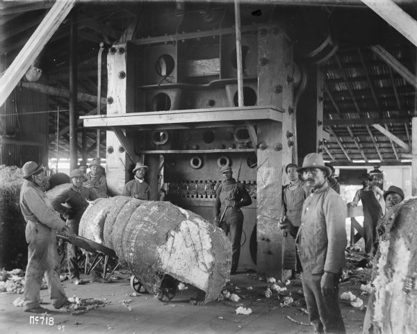 Group of employees, all men or young men, and mostly African American, posing inside manila or cotton processing room of IHC's McCormick Works. One man is holding a cart with a bale of cotton or manila, and two men are standing holding the chains of a large machine. The rest of the group is standing on the left and right. The McCormick Works was built by Cyrus McCormick in 1873 and became part of International Harvester in 1902. The factory was located at Blue Island and Western Avenues in the Chicago subdivision called "Canalport." It was closed in 1961.