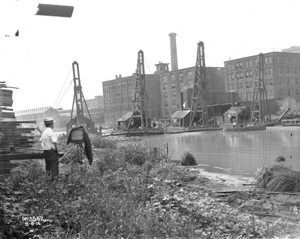 Rear view of company photographer setting up shot of IHC's McCormick Works docks from across the waterfront. Two men are in a long wooden boat, with the name: "Geo. H. Jackson" on it along with "Great Lakes Dredgers." Four rafts, floating in the water and attached to each other with ropes, each have a small building on them with signs that read: "River No. 5. Great Lakes Dredge & Dock Co." Factory buildings and a smokestack are in the background.