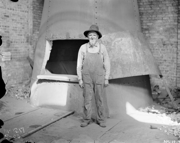 View of worker with a grey beard and hat standing and posing in front of a furnace inside IHC McCormick Works foundry.