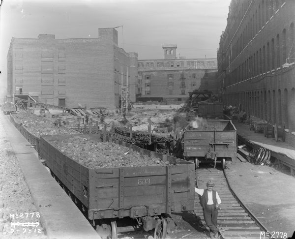 Elevated view of a man standing near railroad cars which appear to be loaded with coal and ore inside IHC's McCormick Works railroad yard. Other men are working in the yard which is full of debris. Scaffolding is seen next to the factory buildings.