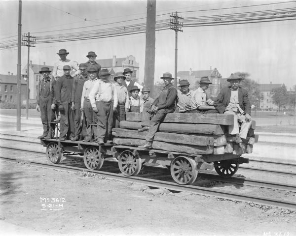 Group portrait of men posing while standing and sitting on two small flat cars linked to each other. One of the cars is loaded with lumber. Another set of railroad tracks is behind the group, and in the background are large buildings in an urban setting. The man on the far left is smoking a pipe, and the man on the far right is smoking a cigar.