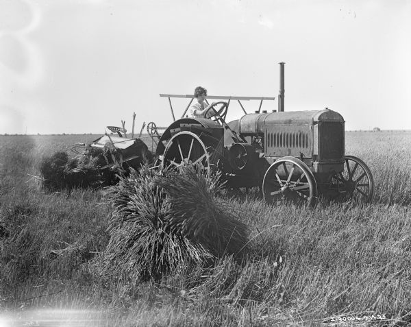Right side view of a woman driving a McCormick-Deering tractor pulling a McCormick-Deering binder. Sheaves of grain are in the foreground.
