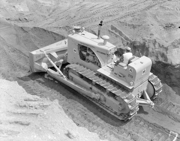 View down slope towards a man driving a crawler tractor to move earth. Some of the decals on the tractor read: "W. Hoggman & Sons Inc.," "Allied" and "TD25." 