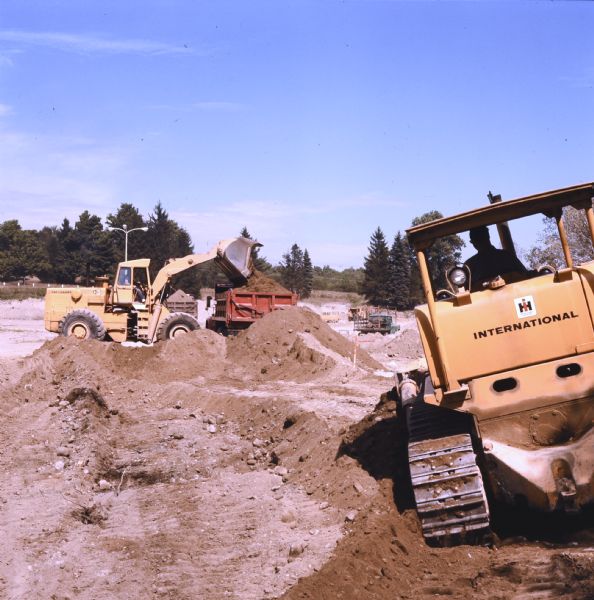 Men moving earth with a TD-20C Crawler Dozer and a H-90E Pay Loader. The payloader is dumping dirt into a dump truck.