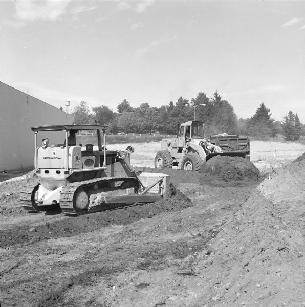 A man is driving a TD-20C Crawler Dozer on the left, and in the background another man is driving an H-90E Pay Loader. They are moving earth near the side of the building. In the far background is a water tower. 