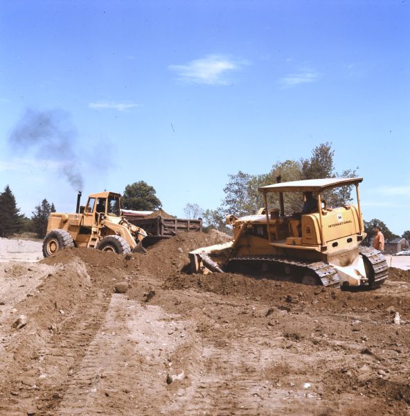 A man is driving a TD-20C Crawler Dozer on the right, and on the left another man is using a H-90E Pay Loader to move earth. Another man is standing and watching on the far right.