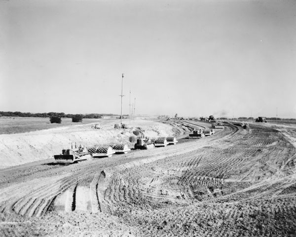 Elevated view of men working with crawler-tractors and sheepsfoot rollers to move earth. Trucks, dump trucks and automobiles are in the background.