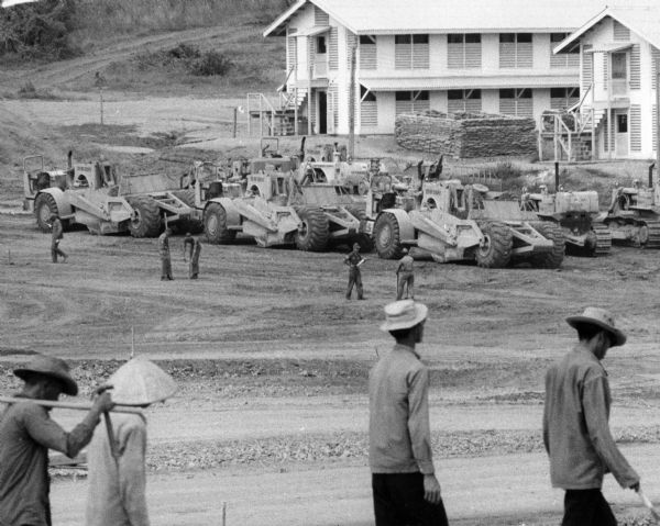 Men are walking in the foreground. In the background more men, in uniform, stand near several International Payscrapers parked in Phan Rang, Vietnam. Recently built barracks are in the background.