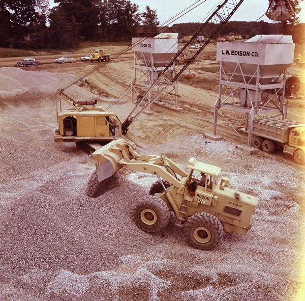 Elevated view of a man operating a Hough Pay Loader with crane and chutes. Trucks and automobiles are parked in the background.