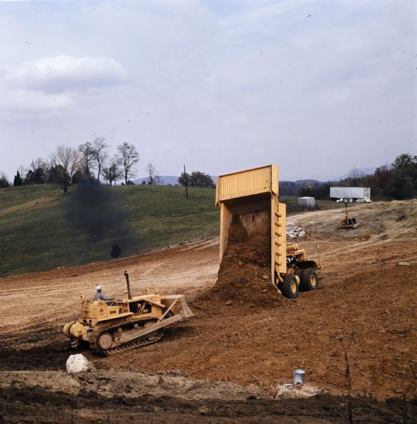 View down hill towards a man operating a TD-20 to move earth that a dump truck is unloading onto the ground.