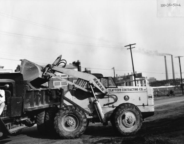 Side view of a Slattery Contracting Company worker using a Hough HM Loader for dumping dirt into the back of truck. Industrial buildings are in the background.